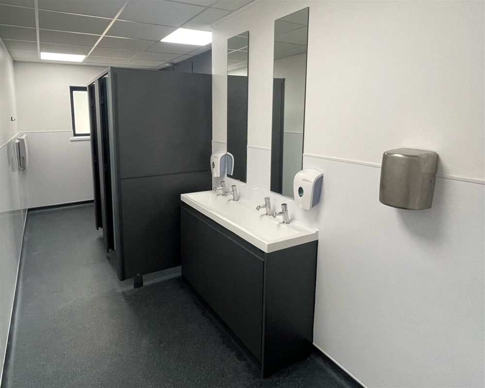 Dark grey vanity unit with countertop washtrough and press action pillar taps and dark grey standard height modular toilet cubicles.